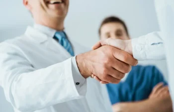Two people in medical attire shake hands, with a third person in scrubs standing in the background, suggesting a professional or collaborative setting.