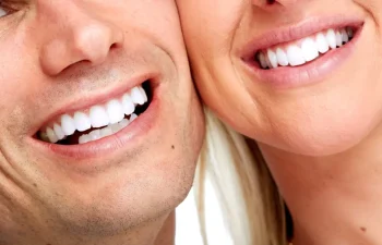Close-up of two people smiling, showing white teeth and partial faces against a plain background.