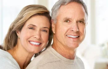 A smiling woman leans on the shoulder of a man, both looking at the camera in a brightly lit indoor setting.