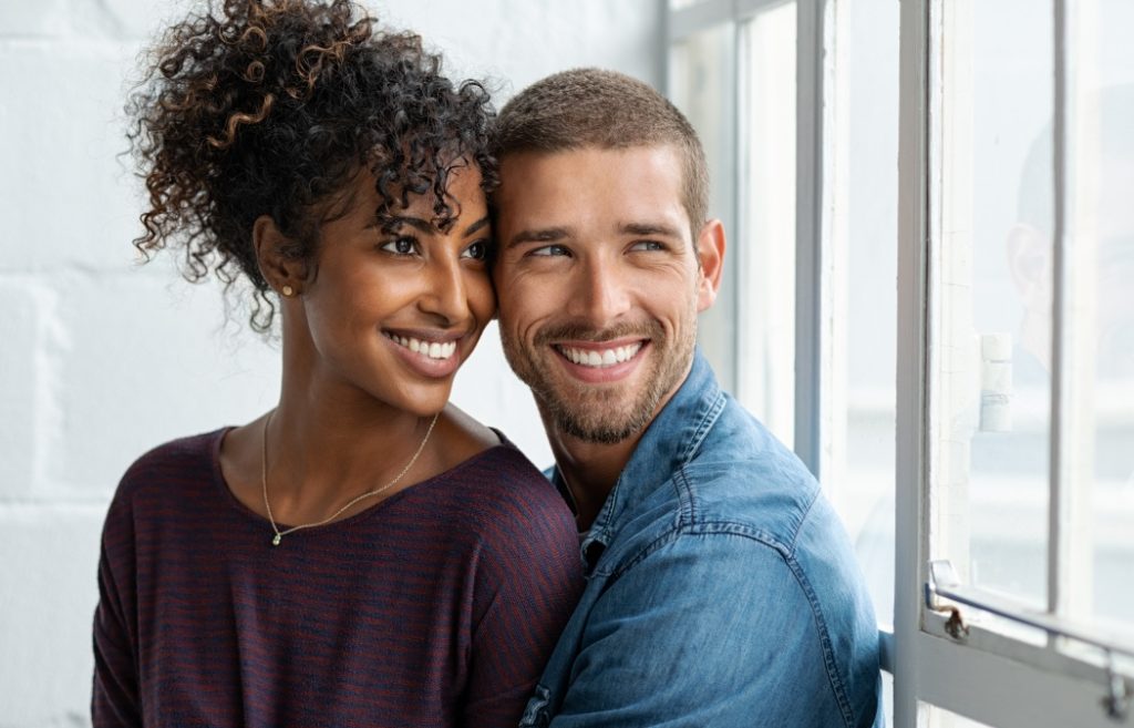 Loving multiethnic couple embracing and sitting near window. Happy girlfriend and smiling boyfriend looking away and thinking about their future together. Young man and african woman contemplating.