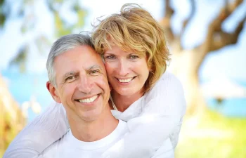 An older couple smiles outdoors, with the woman standing behind the man and embracing him. They are both casually dressed, and trees are visible in the blurred background.
