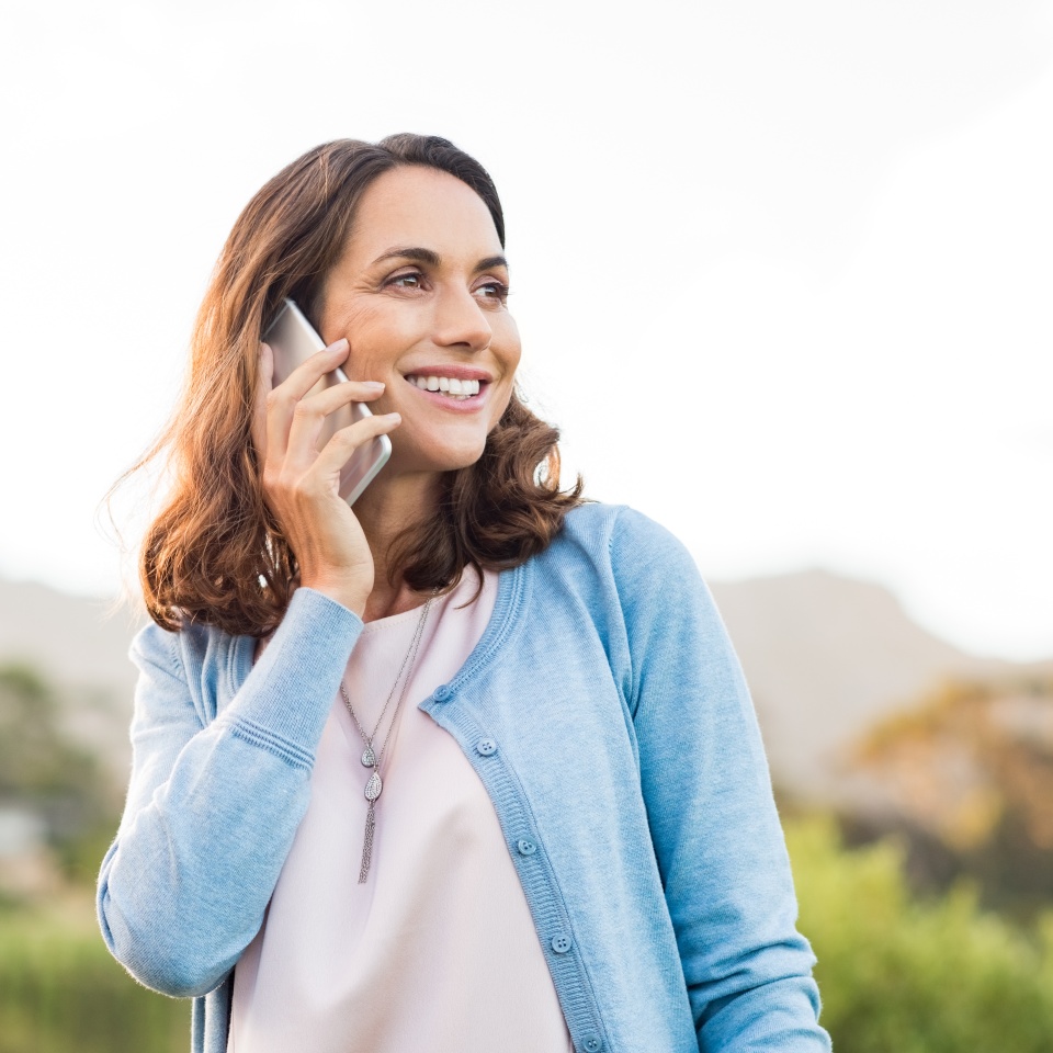 Mature happy woman talking on phone outdoor during sunset. Cheerful hispnic woman using smartphone and looking away. Happy woman in conversation using mobile phone and smiling.