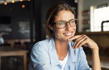 Woman wearing glasses and a light blue shirt sits indoors, smiling and looking to the side, with her hand resting on her chin.