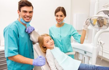 Little girl sitting in the dental chair next dentist and assistant smiling looking at camera