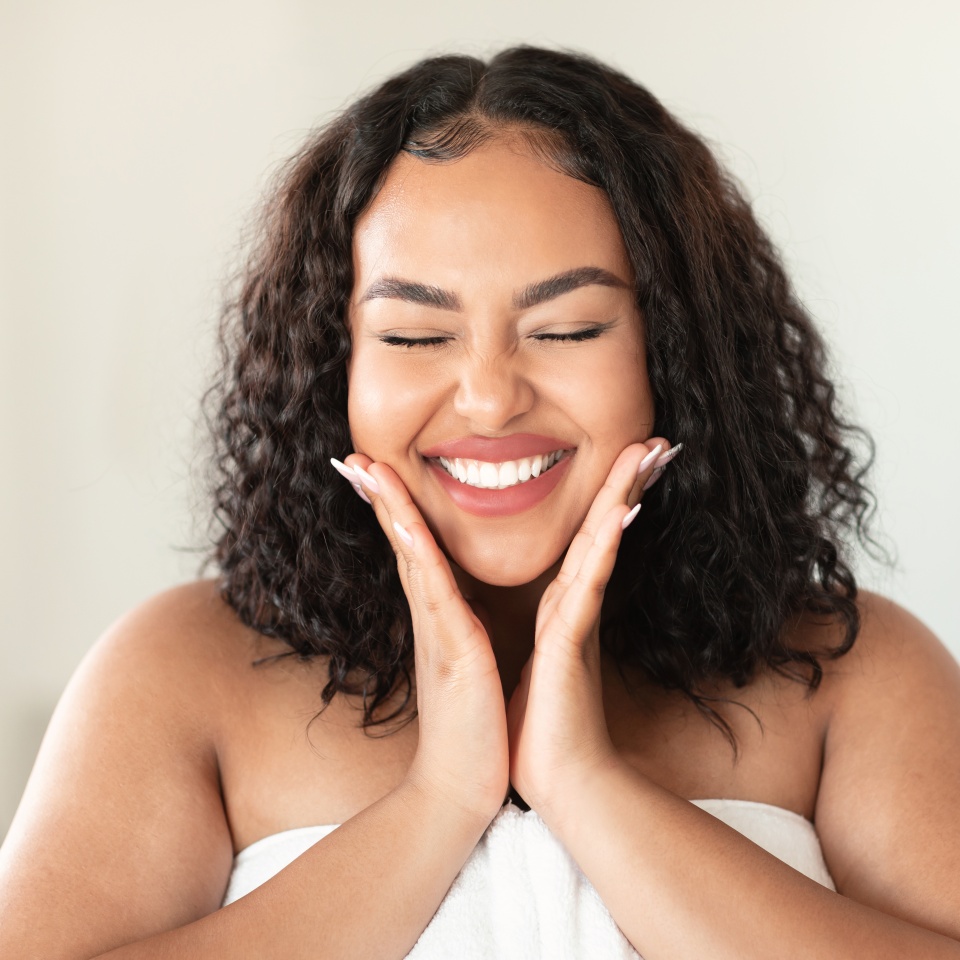 Skin care routine concept. Happy young black plus size woman wrapped in bath towel touching cheeks and smiling