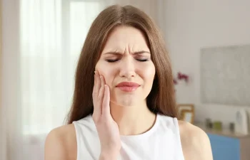 A woman with long brown hair holds her cheek and closes her eyes, appearing to be in pain, possibly due to a toothache.