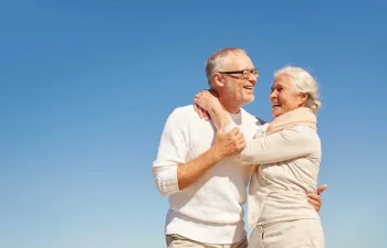 An older couple stands together outdoors under a clear blue sky, smiling and embracing each other.