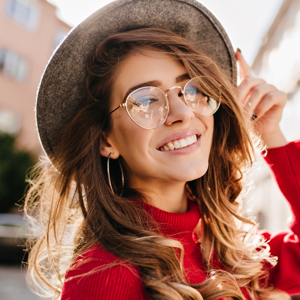 Close-up portrait of cheerful white woman in glasses touching her hat on blur background. Photo of fashionable girl with beautiful brown hair smiling to camera.