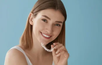 A woman with straight light brown hair smiles while holding a clear dental aligner in her hand against a light blue background.