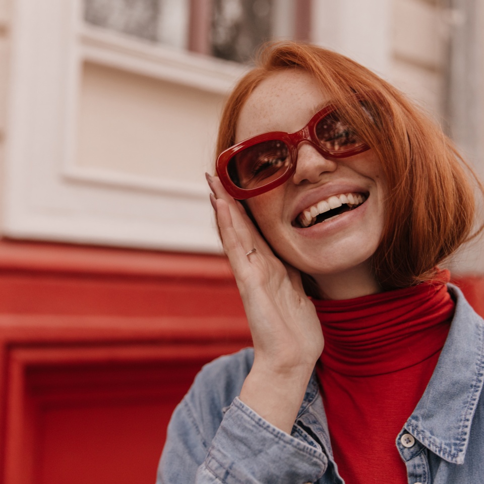 Close-up portrait of young beautiful lady posing outdoors. Pretty red-haired girl wearing turtleneck and blue shirt, adjusting trendy sunglasses and smiling against bright wall background