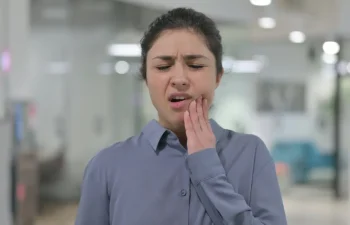 A woman touches her cheek and winces in pain, possibly experiencing a toothache or jaw discomfort, in an indoor setting.