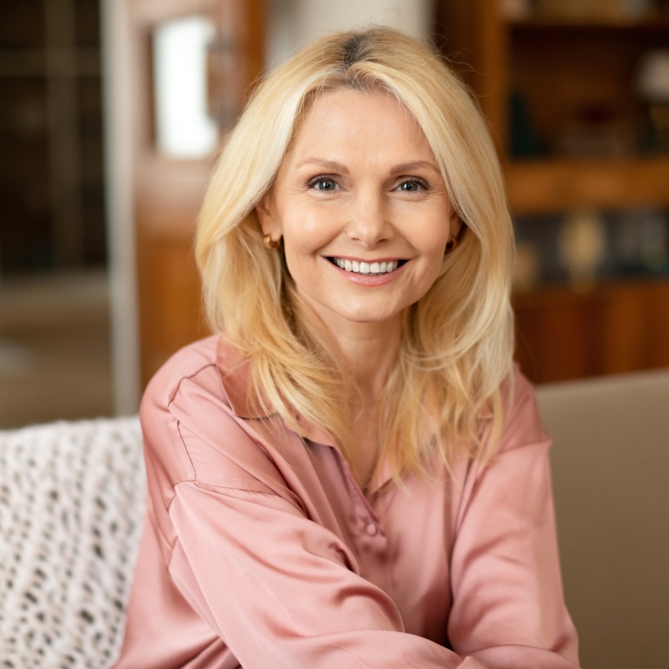 Portrait Of Blonde European Mature Woman Posing Smiling To Camera Sitting On Sofa At Home. Lady Resting On Cozy Couch In Modern Living Room Indoor. Ageless Beauty And Elegance Concept