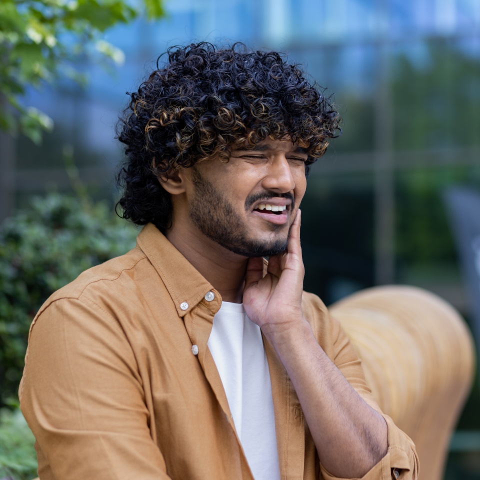 A young hispanic man sits outside on a bench, grimacing and holding a hand to his cheek. Suffering from toothache. Close-up photo.