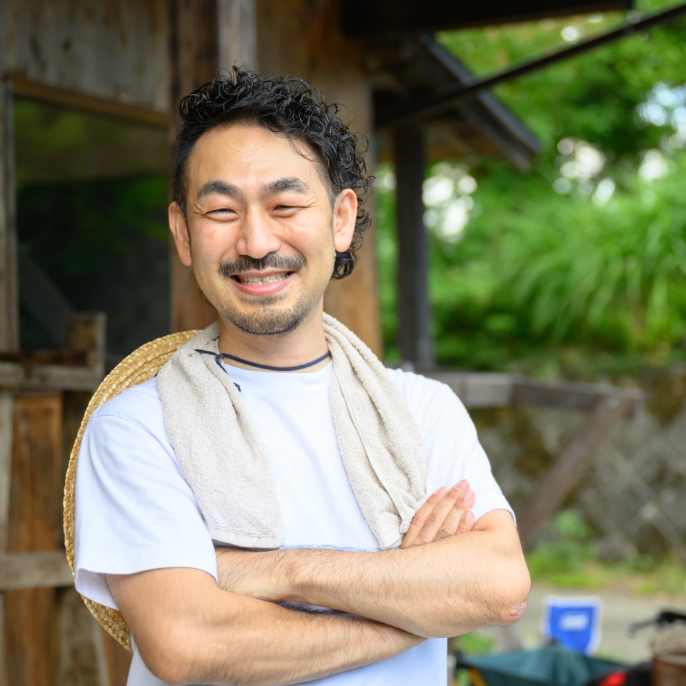 A Japanese farmer stands in the yard of an old house in the Japanese countryside with his arms folded and a smile on his face. The photo shows the upper half of his body, looking at the camera.