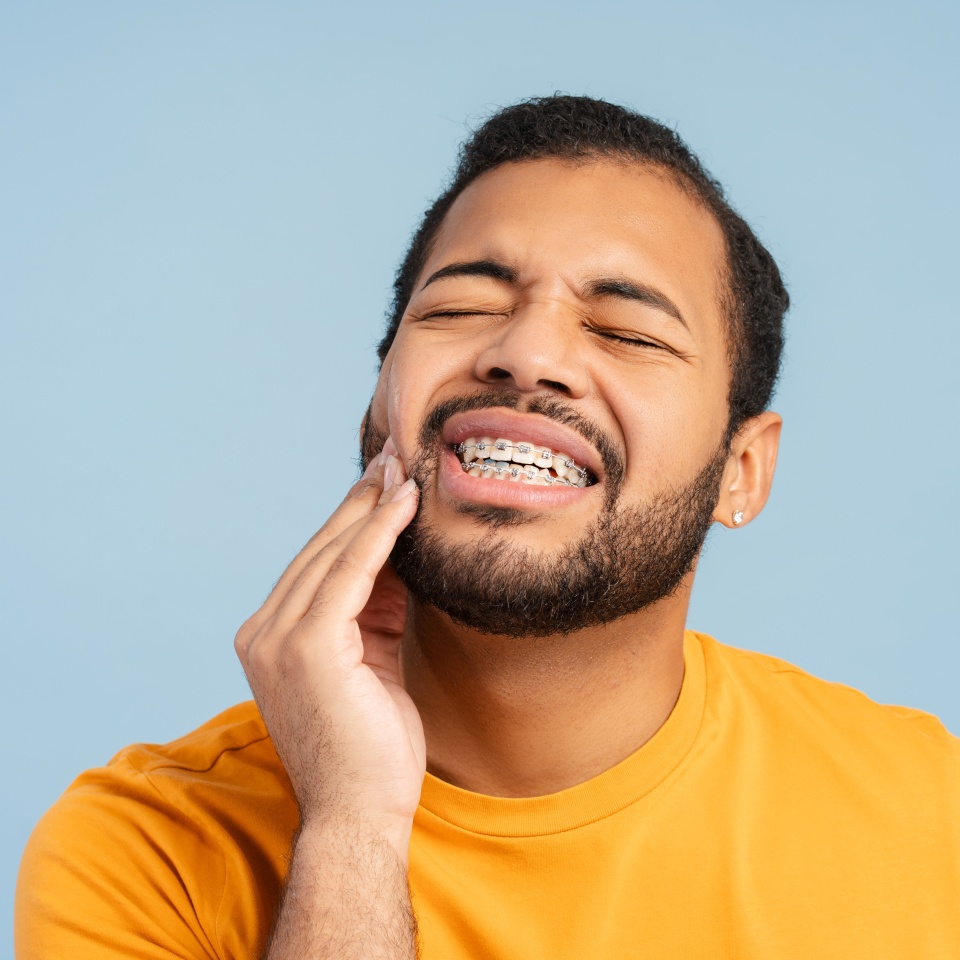 Portrait of troubled, african american man with braces, holding his cheek in pain, against a blue background. Illustrates dental discomfort concept