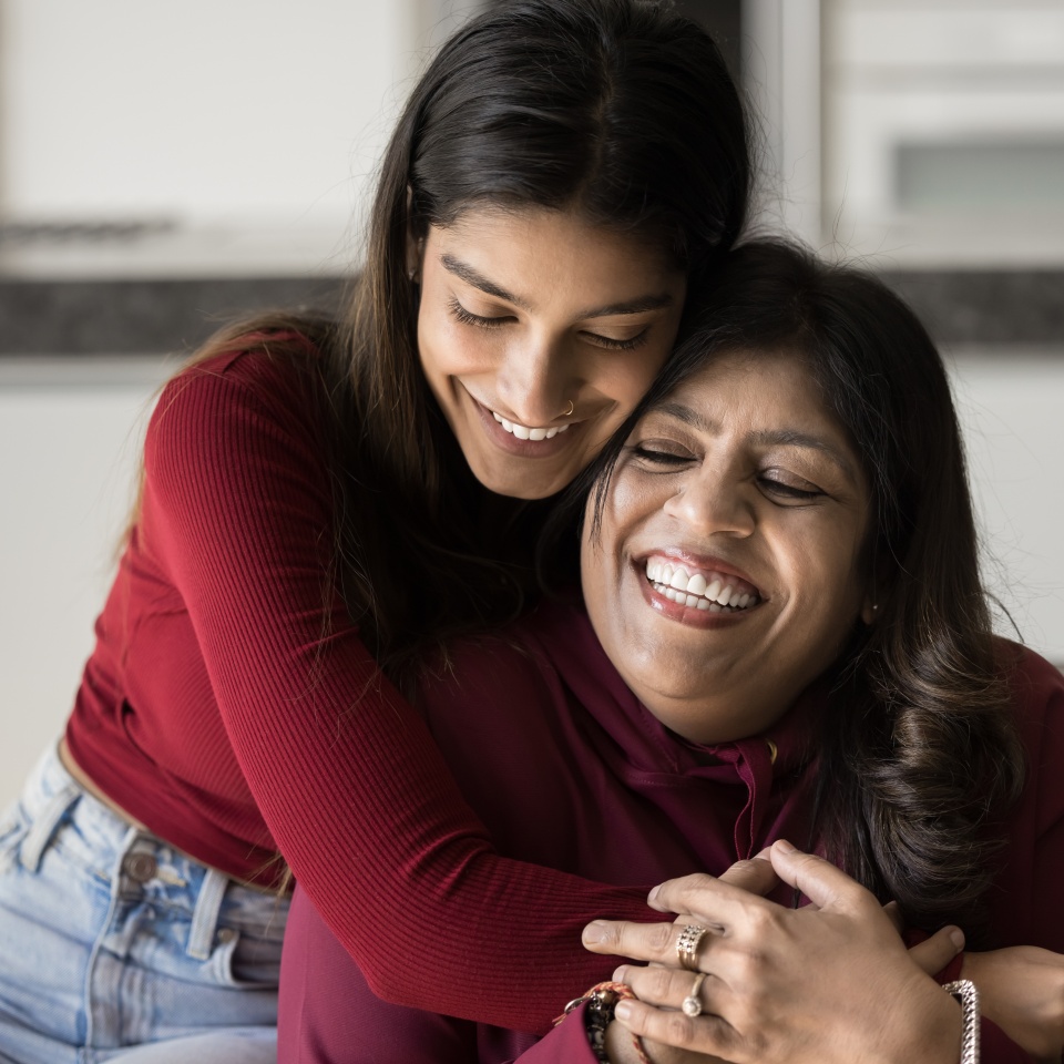 Happy beautiful young Indian daughter woman embracing senior mom from behind with love, tenderness. Cheerful mother and adult kid hugging with closed eyes on home couch, smiling, laughing