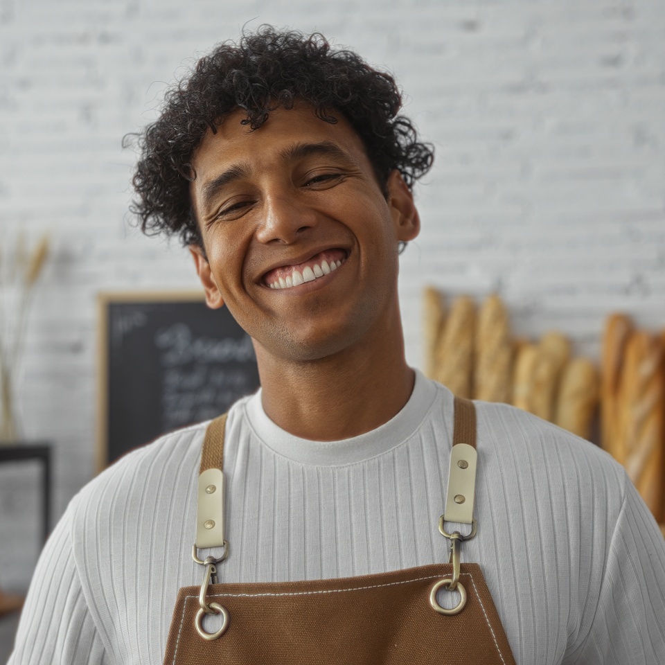 Young hispanic man smiling in a bakery shop interior with shelves of baguettes in the background