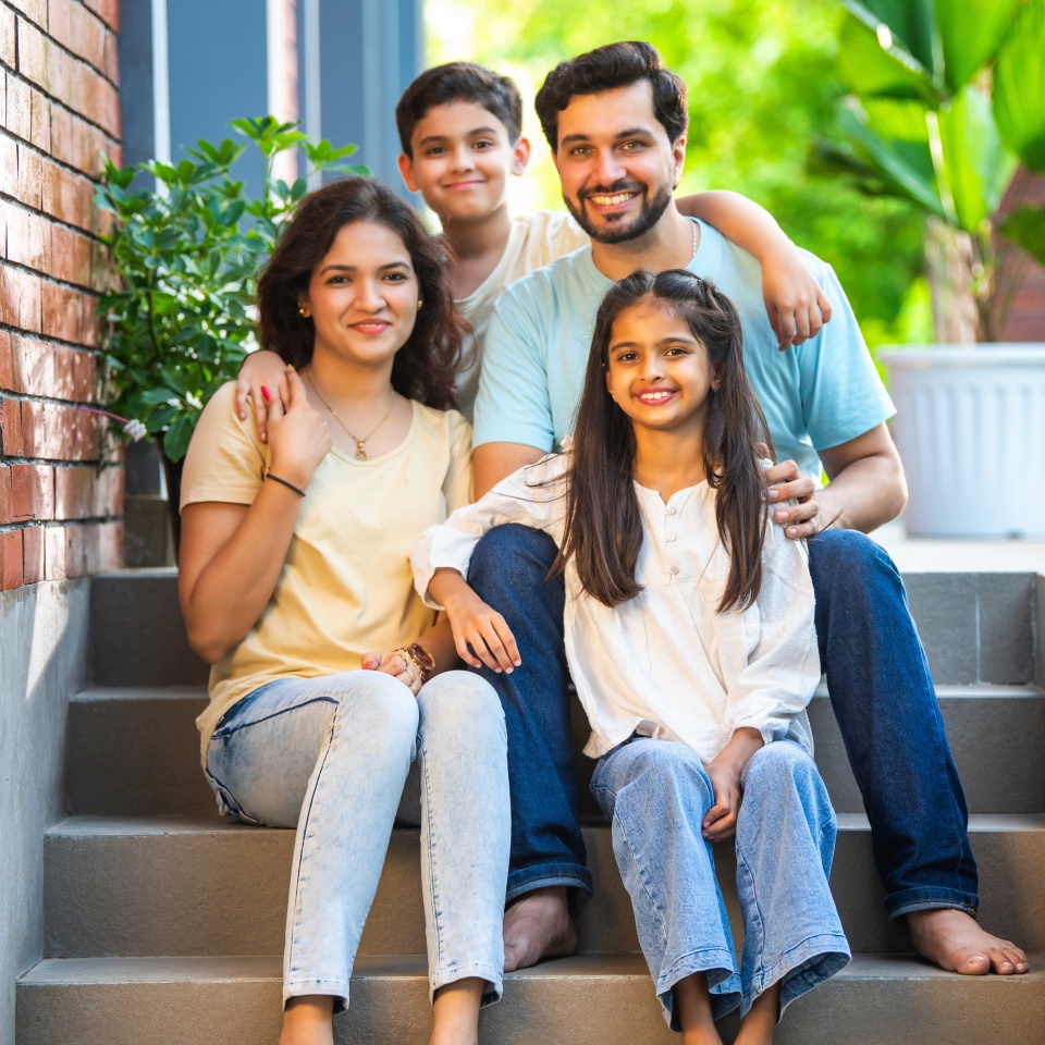 Portrait of Smiling Indian family of four, young parents and two kids, sitting joyfully on the front steps of their home, sharing a warm and happy moment together, celebrating love, family bond