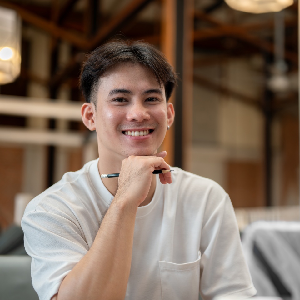 A handsome, happy Asian man sitting at a couch in a coffee shop with his friends, resting his chin on his hand and smiling at the camera. people and lifestyle concepts