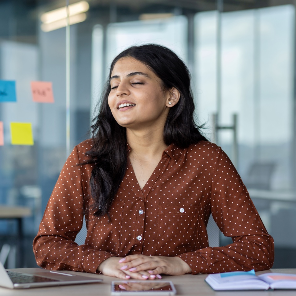 Successful woman relaxing and meditating with closed eyes, breathing exercises at workplace inside office with notebook. Businesswoman smiling dreaming and visualizing future results of achievement.