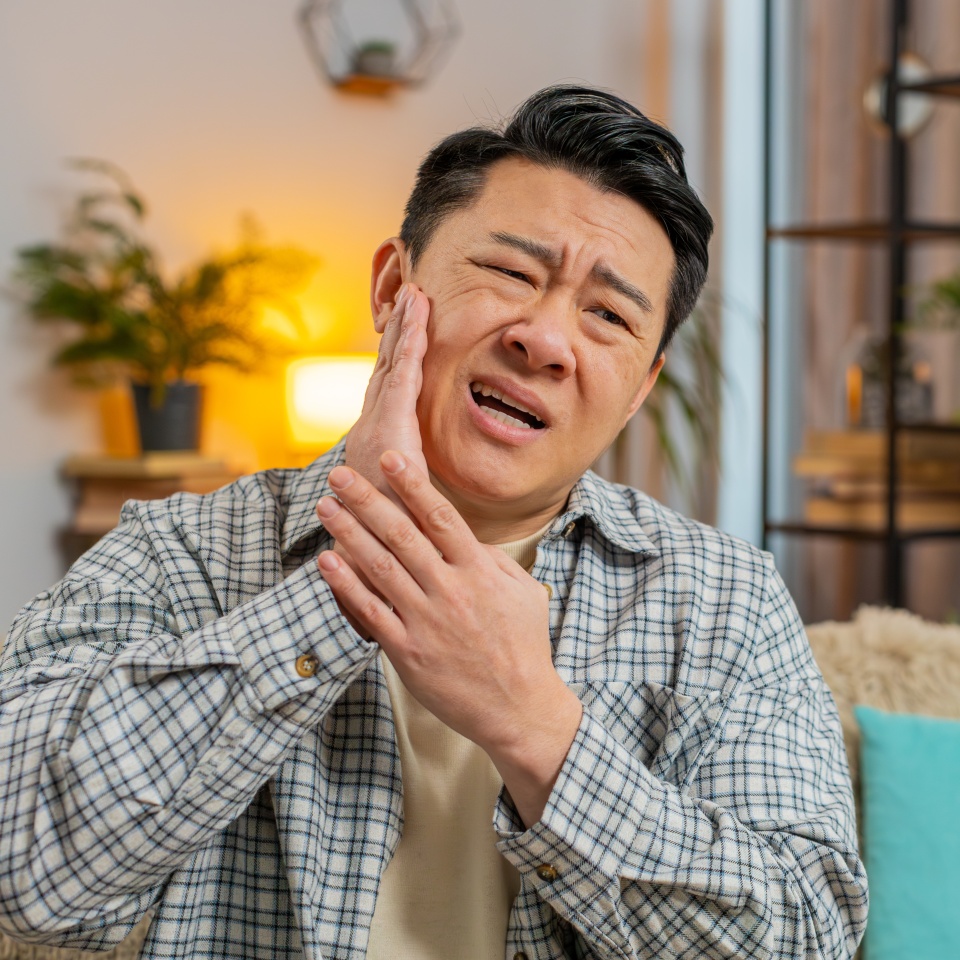 Dental problems. Young man touching cheek, closing eyes with expression of terrible suffer from painful toothache, sensitive teeth, cavities. Chinese guy at home apartment living room on cozy sofa.