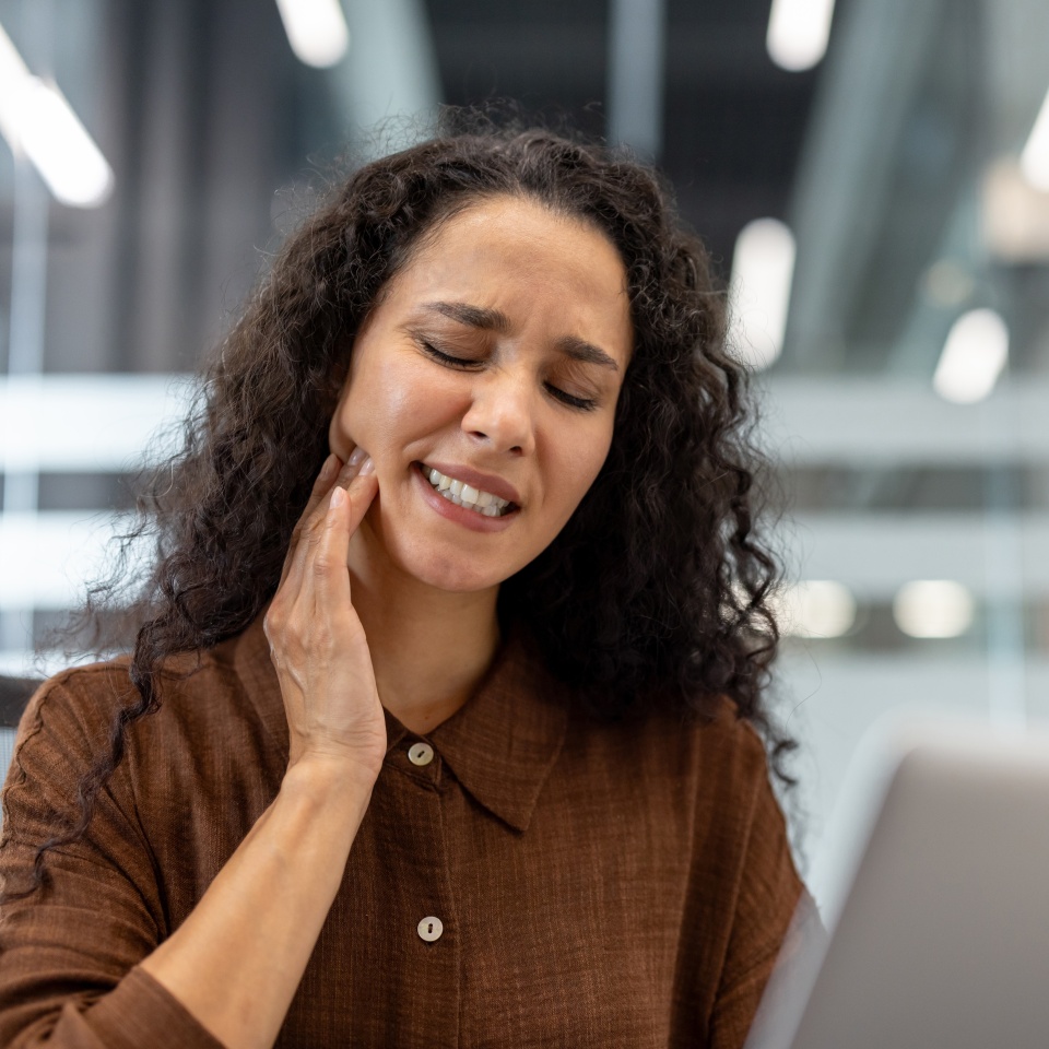 Woman suffering from a painful toothache, holding her jaw and closing her eyes in discomfort, struggling to concentrate on working with her laptop at her office desk