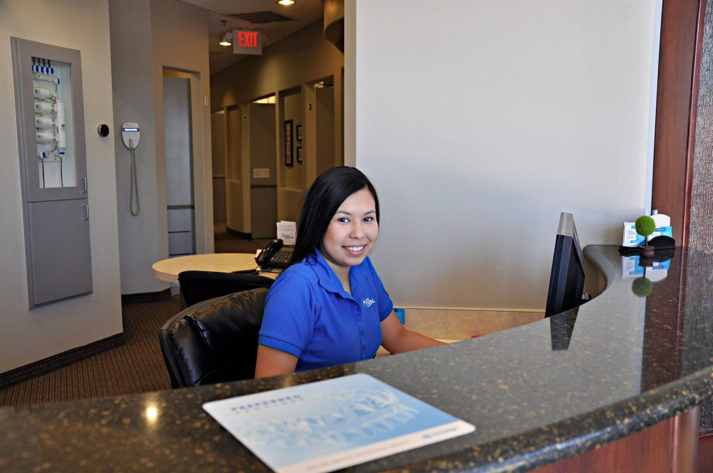 A woman in a blue shirt sits at a reception desk, smiling at the camera, in a modern office lobby with hallways and office doors in the background.
