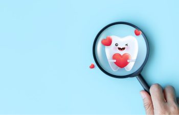 A hand holds a magnifying glass over a cartoon tooth with a smiling face, surrounded by red hearts, on a blue background.