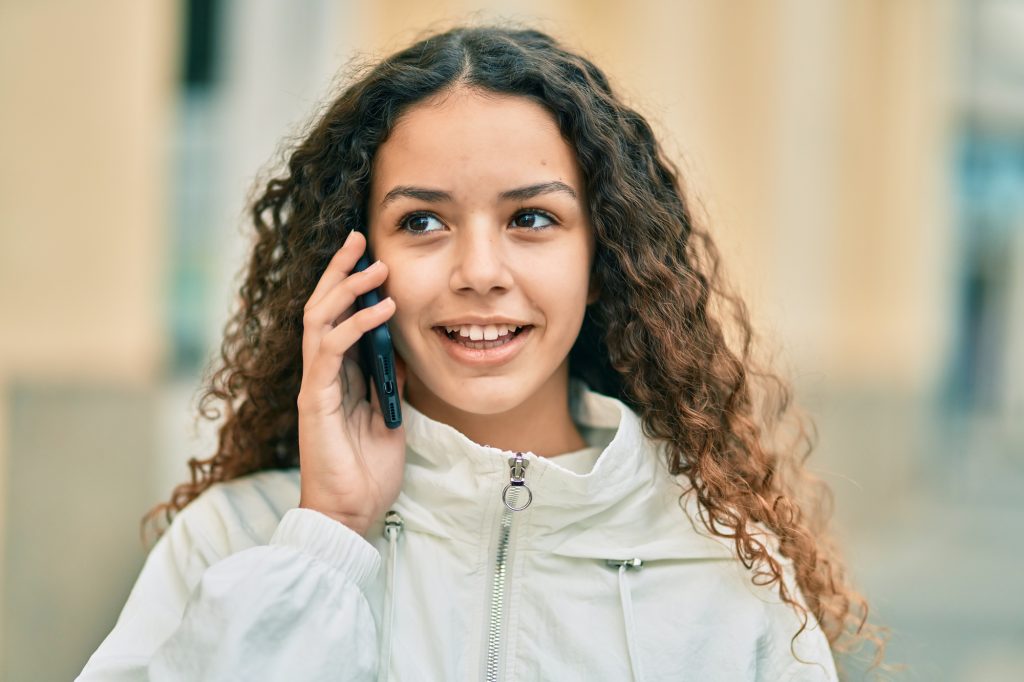 A young person with curly hair and a white jacket is smiling while talking on a smartphone outdoors.