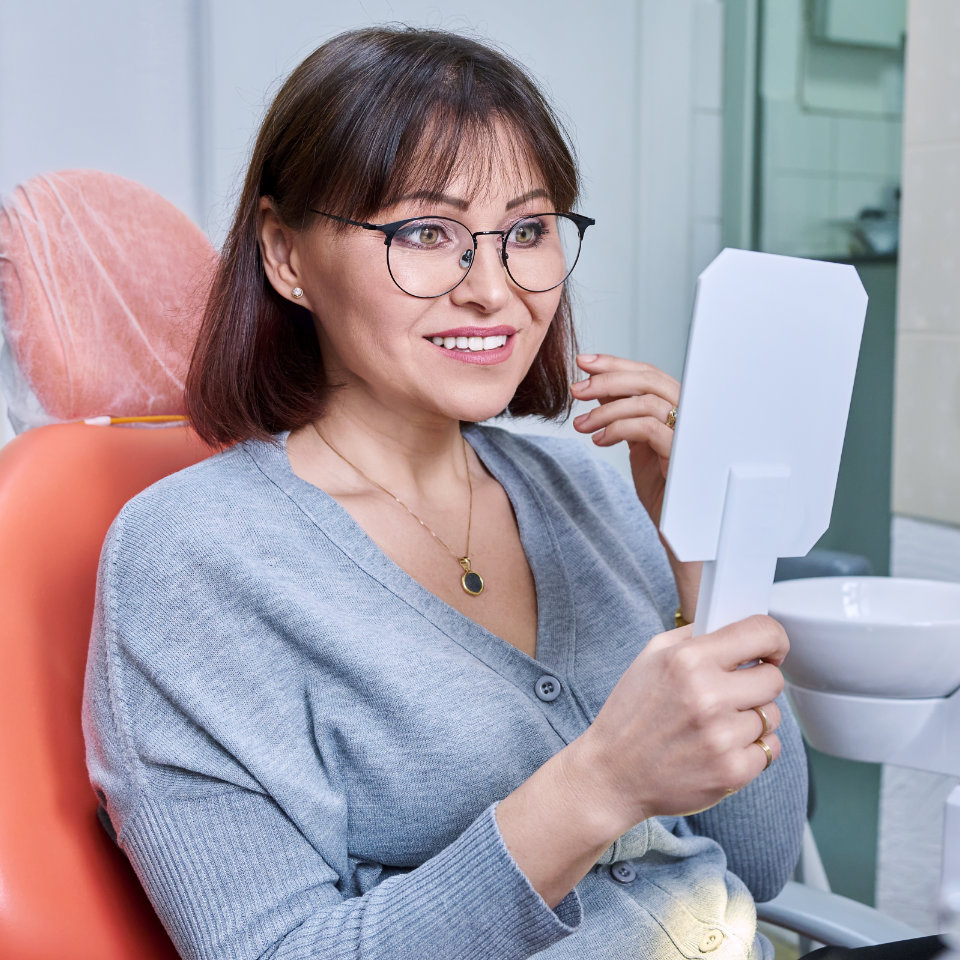 A woman sitting in a dental chair holds a mirror and smiles, seemingly inspecting her teeth.
