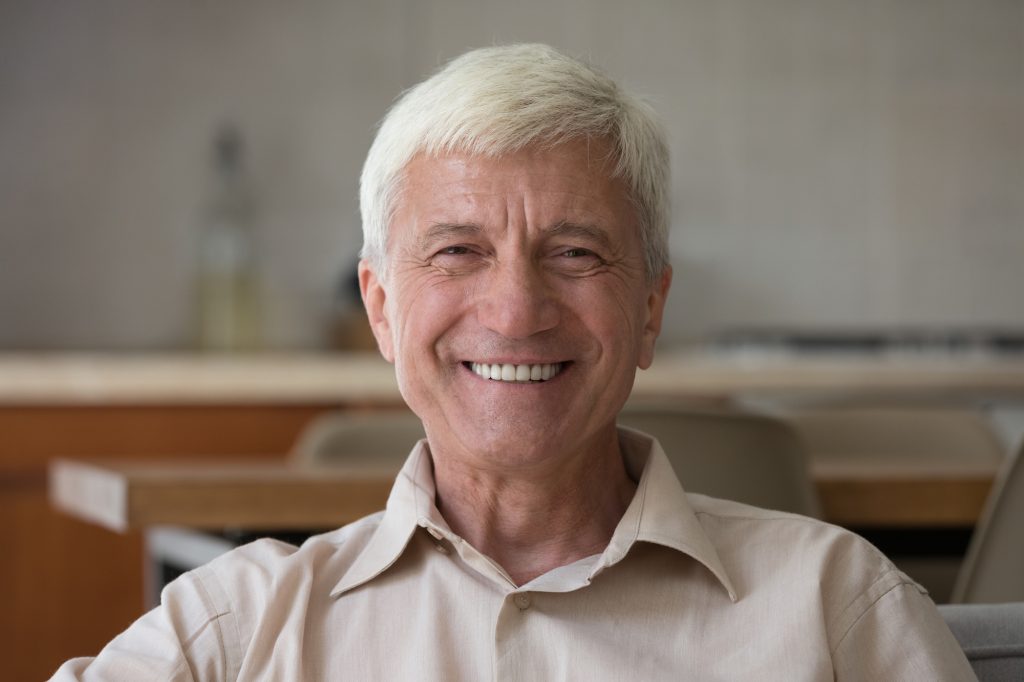 Older man with short white hair and a beige shirt smiles while sitting indoors, with a kitchen in the blurred background.