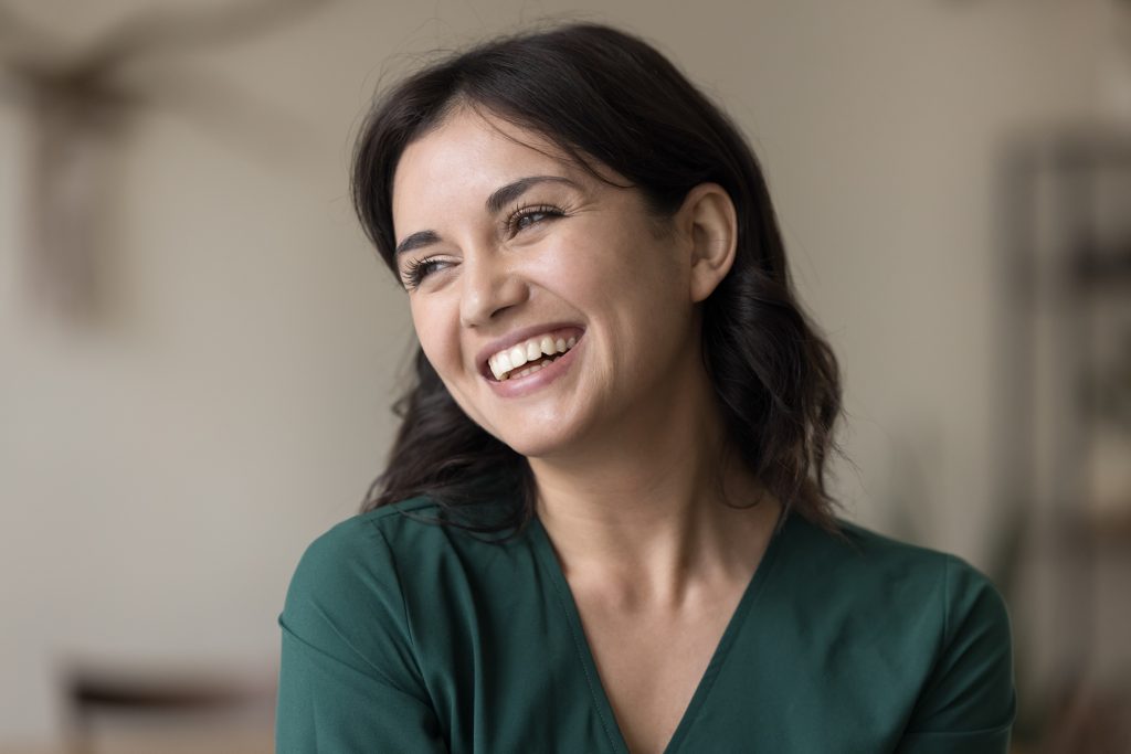 Woman with dark hair wearing a green shirt smiles and looks slightly to the side in a softly lit indoor setting.