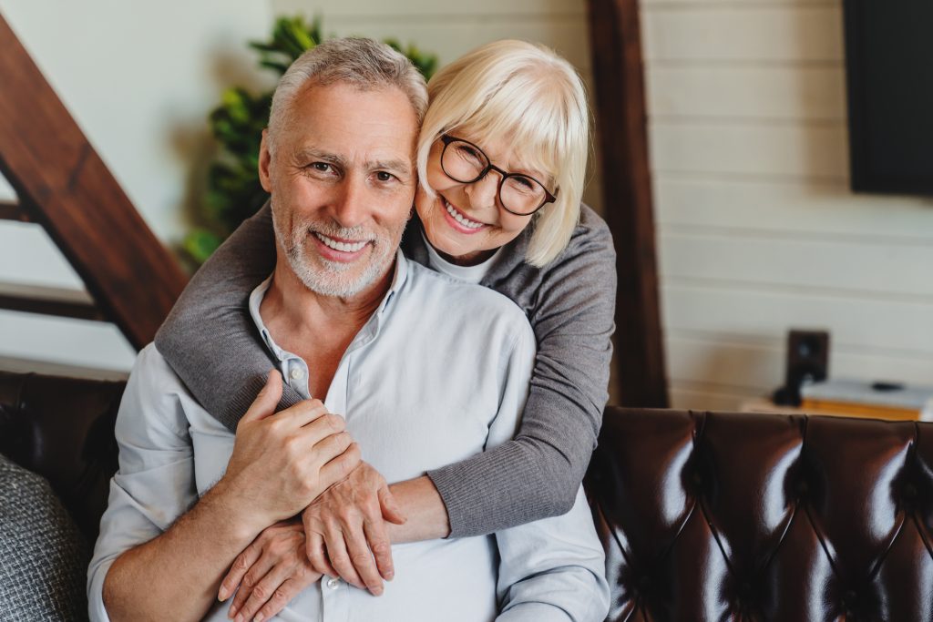 An older woman with glasses smiles and hugs an older man from behind as they sit together on a brown leather couch in a cozy, well-lit living room.