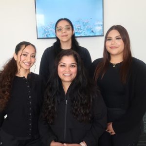 Four Active Dental team members pose together indoors, smiling and wearing coordinated black uniforms. They are standing in a bright office space with a wall-mounted screen behind them.