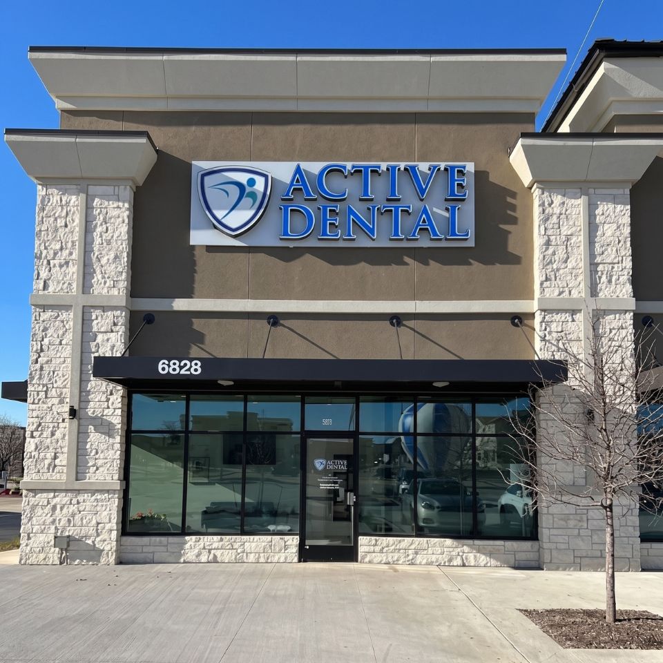 A daytime, front-facing shot of the exterior of "Active Dental," located in a modern commercial strip building. The storefront features large glass windows and a glass door, reflecting cars and a building opposite. Above the black awning, which displays the address number "6828," is a large sign with blue lettering reading "ACTIVE DENTAL" next to a blue shield logo. The building's facade consists of smooth beige stucco walls flanked by decorative white stone pillars, under a clear blue sky.