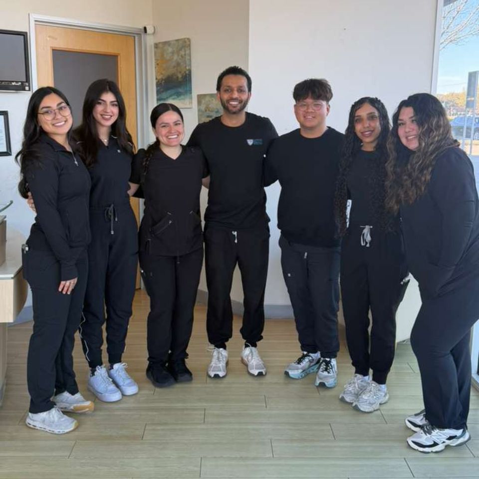 A group of seven smiling Active Dental team members standing together in a semi-circle inside the clinic. They are all wearing professional matching black scrubs and sneakers, posing happily for a team photo in a brightly lit office area.