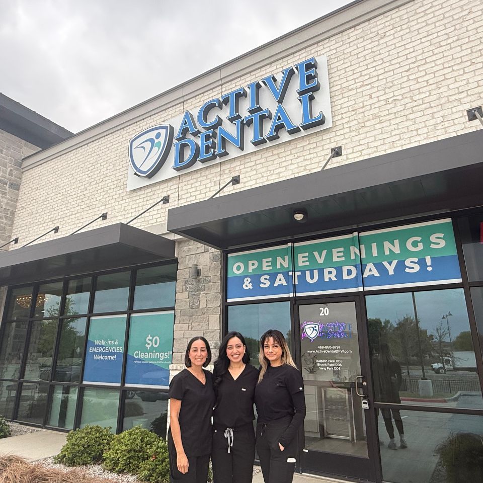 Three smiling female staff members in black medical scrubs standing outside the entrance of an Active Dental clinic. The storefront features a white brick facade, large glass windows with signs reading "Walk-ins & Emergencies Welcome!" and "$0 Cleanings!", and a prominent banner over the door that says "Open Evenings & Saturdays!" The blue Active Dental sign with its shield logo is mounted above the entrance.