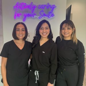 Three smiling female Active Dental staff members wearing black medical scrubs standing side by side indoors. Behind them, a bright purple neon sign on the wall reads "Actively caring for your Smile" in cursive lettering.