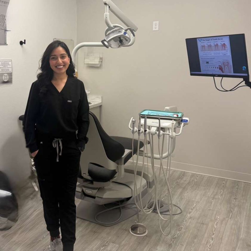 A smiling female dental professional in black scrubs standing inside a modern treatment room at Active Dental. The room is equipped with a grey patient chair, overhead dental light, a tool delivery system, and a wall-mounted monitor displaying an educational chart about tooth decay.
