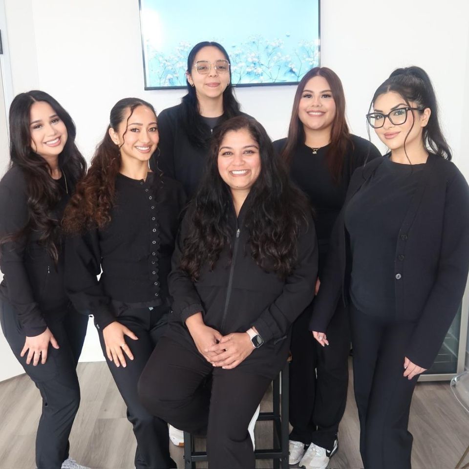 Six Active Dental team members pose together indoors, smiling and wearing coordinated black uniforms. They are gathered in a bright office space with a wall-mounted screen behind them.