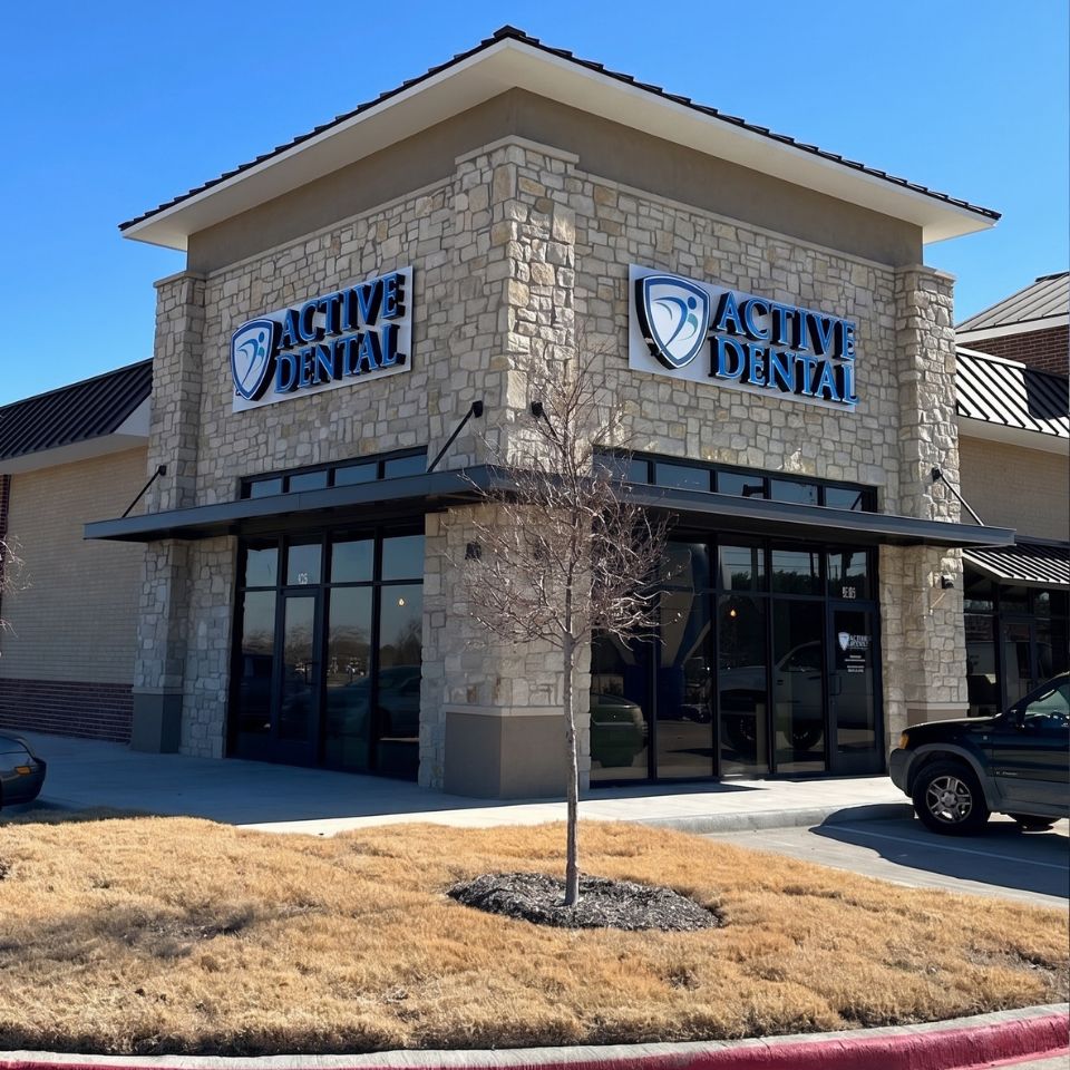 Exterior view of an Active Dental clinic in a stone-front building on a sunny day, with the practice’s logo and signage displayed above the windows. A small tree, sidewalk, parked vehicle, and clear blue sky are visible in front of the office.