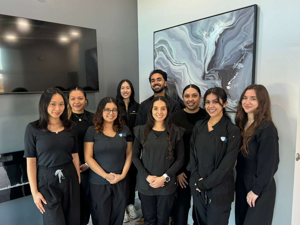 Active Dental team members pose together in a modern office, wearing coordinated black uniforms and smiling at the camera. The group stands in front of a gray wall with a large abstract artwork and a wall-mounted screen in the background.