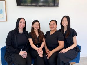 Four Active Dental team members sit together on a blue couch in a bright office, smiling and wearing coordinated black uniforms. A wall-mounted screen and framed artwork are visible behind them.