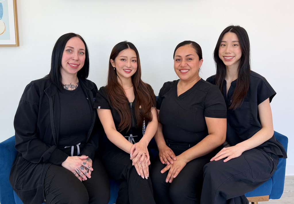 Four smiling female Active Dental staff members, wearing professional black medical scrubs, sitting side by side on a blue sofa in a brightly lit waiting area.