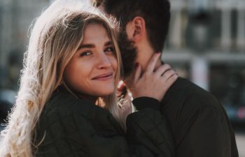 Close up portrait of charming girl looking at camera and smiling while cuddling with bearded man on the street