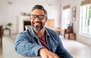 Happy mature middle eastern man wearing eyeglasses with beard sitting on couch at home. Portrait of indian man relaxing at home and looking away with big smile. Handsome mid adult guy with specs thinking about his future.